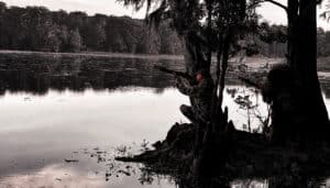 Two people in camouflage gear are hunting by the edge of a body of water surrounded by trees, aiming rifles in the direction of the water.