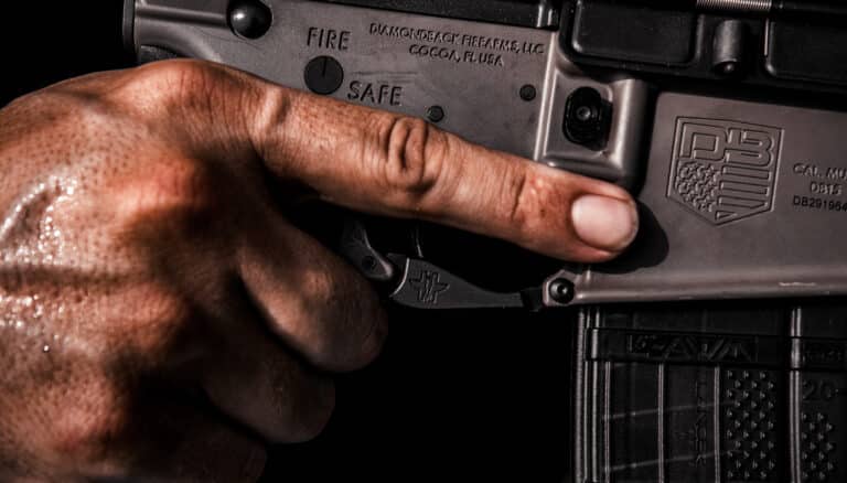 A close-up of a person's hand with visible drops of sweat, pressing the safety switch on a firearm marked with "FIRE" and "SAFE". The gun also features various engraved text and logos.