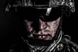 A close-up of a soldier wearing camouflage helmet, dark sunglasses, and a scarf, with a serious expression and dirt on their face.