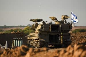 Israeli military vehicle with surveillance equipment and an Israeli flag stationed in a dirt area, with artillery shells nearby and open landscape in the background.